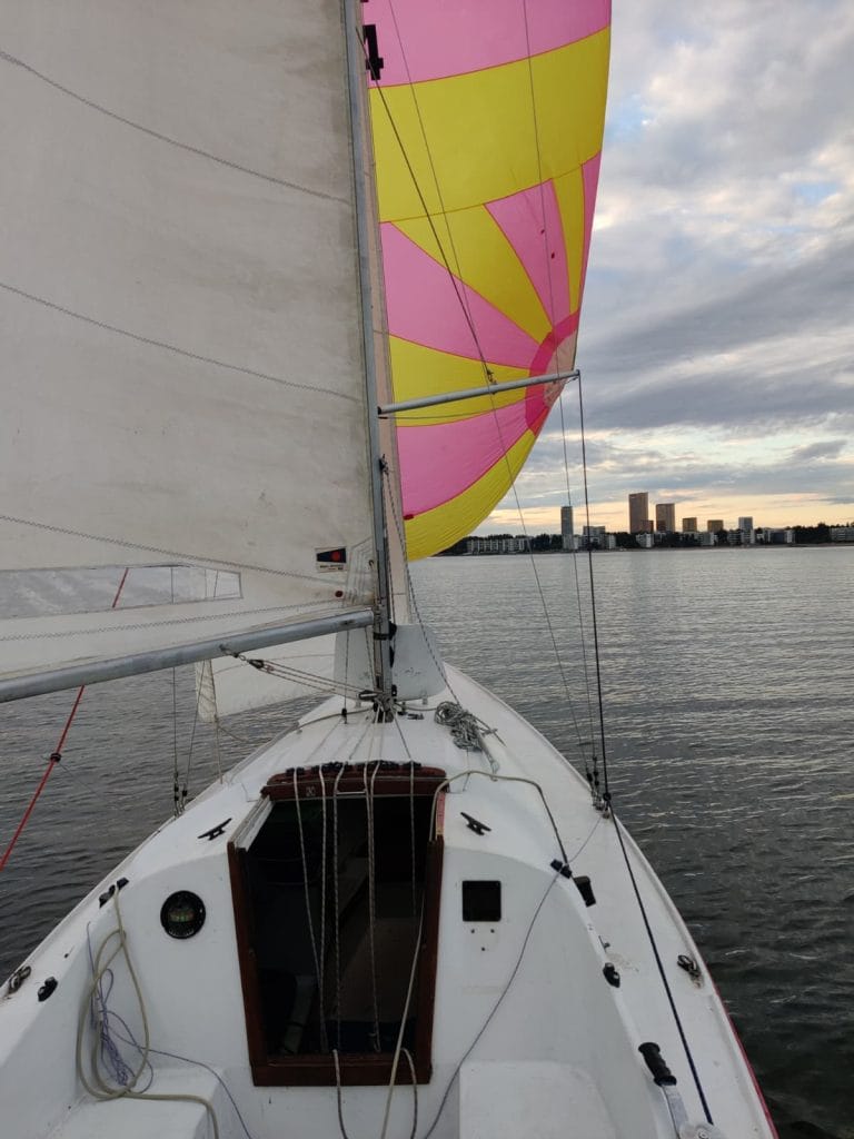 Kuva H-veneen kyydistä jossa värikäs spinaakkeri ylhäällä ja taustalla ilta-aurinkoinen Aurinkolahden ranta.